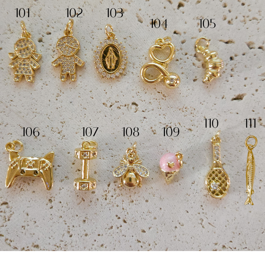 Twelve small gold charms or pendants are arranged on a light stone background, each labeled with a number from 101 to 112. The charms include animal and human-like shapes, a heart with a pink stone, a fish, and other decorative motifs.
