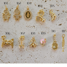 Twelve small gold charms or pendants are arranged on a light stone background, each labeled with a number from 101 to 112. The charms include animal and human-like shapes, a heart with a pink stone, a fish, and other decorative motifs.